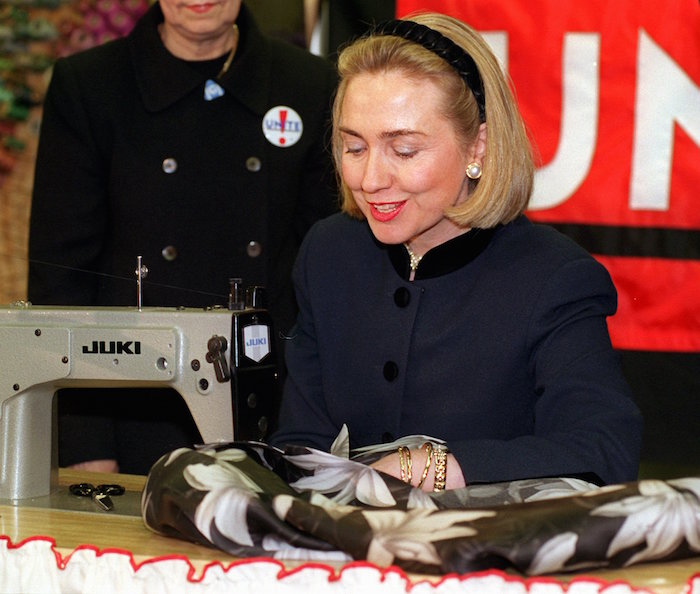 In this March 6, 1996, file photo, then-first lady Hillary Rodham Clinton sews a label into a womans floral print jacket at designer Nicole Millers New York showroom as the symbolic first label of UNITE, the organization that resulted from the merger of the International Ladies Garment Workers Union and the Amalgamated Clothing and Textile Workers Union. Clinton’s support for trade deals has fluctuated with the political calendar. As first lady, she trumpeted the North American deal brokered by her husband, telling unionized garment workers in 1996 that the agreement was proving its worth. Now, early in her Democratic presidential campaign, she’s striking a different tone.