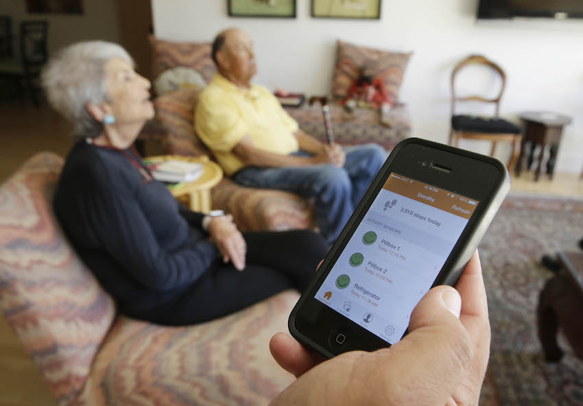 In this March 17, 2015 photo, Phil Dworsky shows off an app on his phone that displays the movements of his parents, Dorothy, 80, left, and Bill Dworsky, 81, rear, at their home in San Francisco. Each time an elder Dworksy opens the refrigerator, closes the bathroom door or lifts the lid on a pill container, tiny sensors in their home make notes on a digital logbook, which the younger Dworsky monitors daily on his smartphone. 
